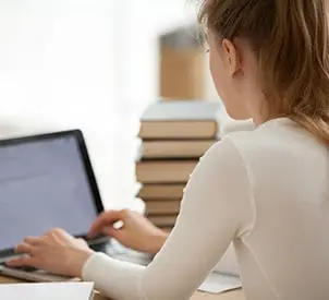 Close-up of a dedicated woman student sitting at a desk, typing and writing a message or email while looking at a computer with a mock-up blank screen, viewed over the shoulder. The girl is surrounded by heaps of books, engaged in studying at home or in the library. Ideal for those pursuing top-notch dissertation coach and academic excellence.